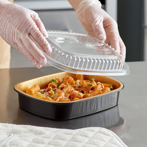 A person in gloves placing a plastic lid on a ChoiceHD black and gold foil tray filled with pasta and sauce.
