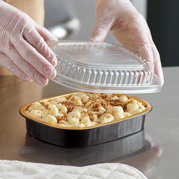 A person in gloves holding a ChoiceHD black and gold foil entree pan with a plastic lid over macaroni and cheese.
