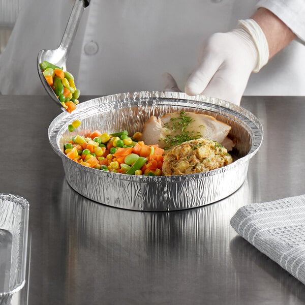A hand in a white glove spooning vegetables into a Choice 9" round foil take-out pan.
