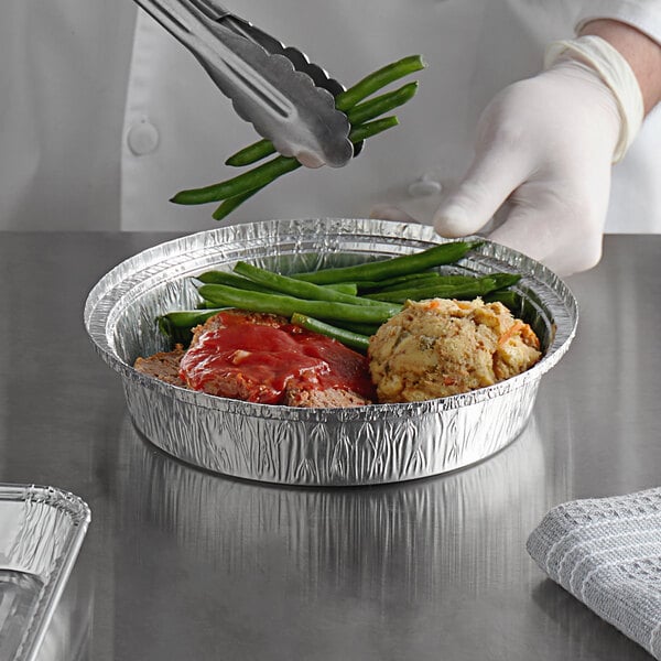 A chef using tongs to pick up food from a Choice foil take-out container.