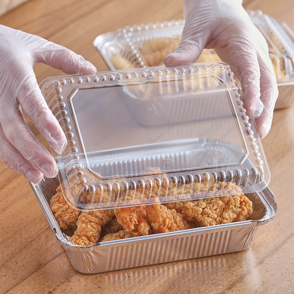 A hand in a plastic glove holding a clear plastic lid over a container of chicken.