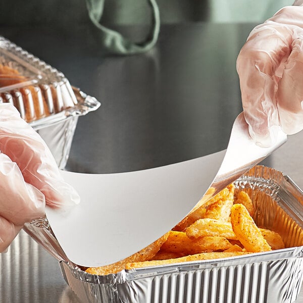 A person in gloves holding a tray of food covered with a foil-laminated board lid.