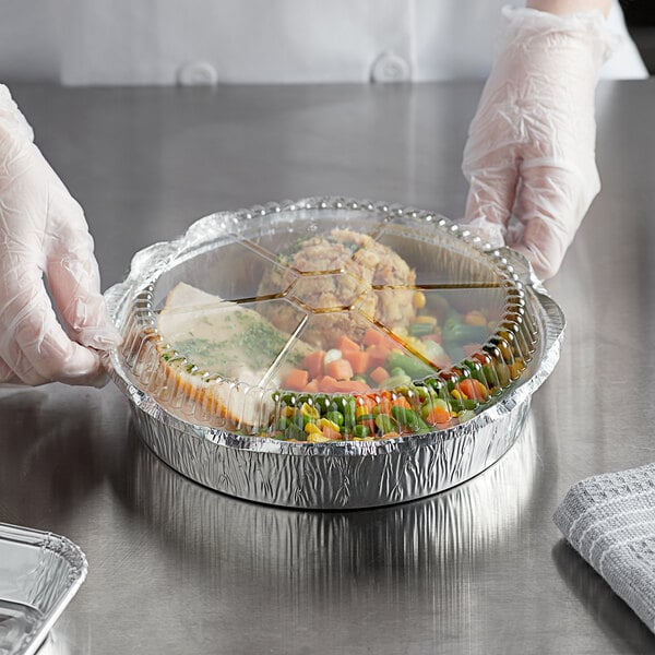 A person in gloves holding a tray of food in a Choice 9" round foil take-out pan with a dome lid.