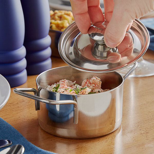A hand opens the lid to a Vollrath mini stainless steel casserole dish filled with shrimp and herbs.