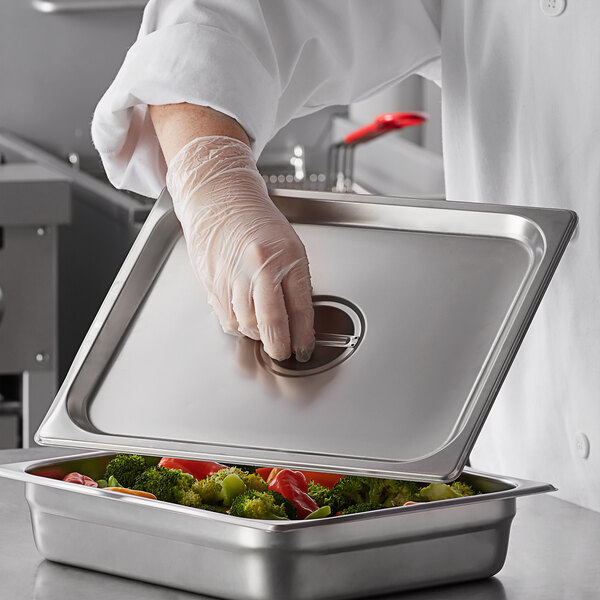 A person in gloves holding a Carlisle stainless steel hotel pan cover over a metal tray of food.