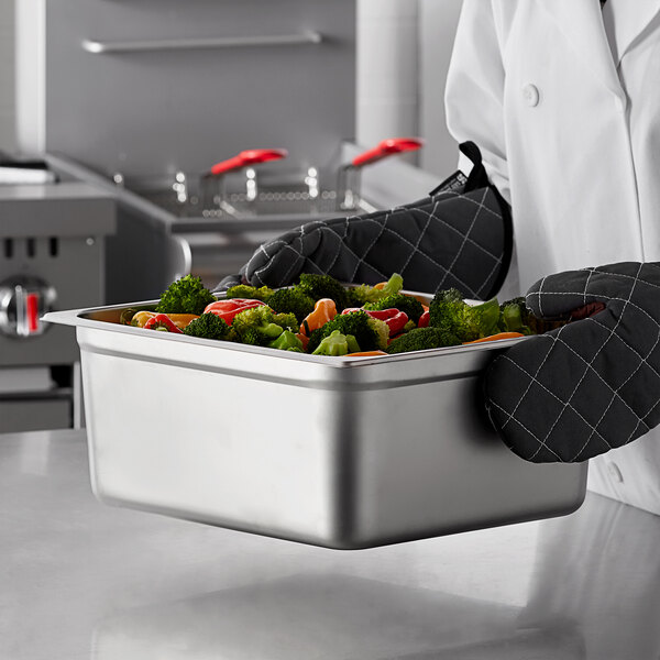 A chef holding a Carlisle stainless steel steam table pan filled with broccoli and tomatoes.