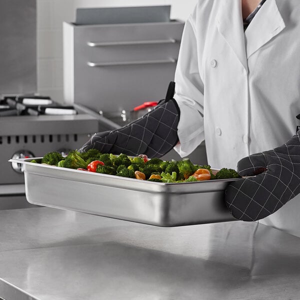 A chef in a black apron holding a Carlisle stainless steel steam table pan full of vegetables.
