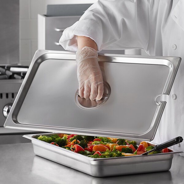 A person wearing a white coat and gloves using a Carlisle stainless steel slotted pan cover to serve vegetables.