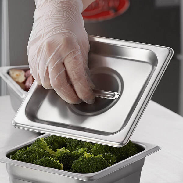 A hand in a glove holding a Carlisle stainless steel tray with a lid of broccoli.