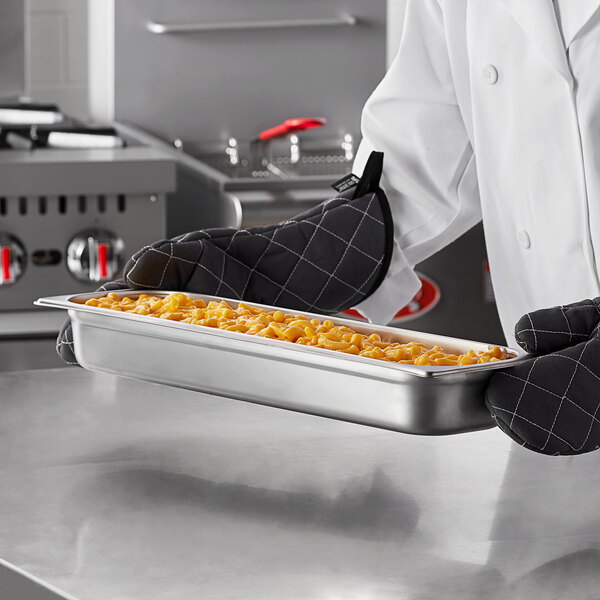 A chef in a black apron holding a white Carlisle stainless steel tray of macaroni and cheese.