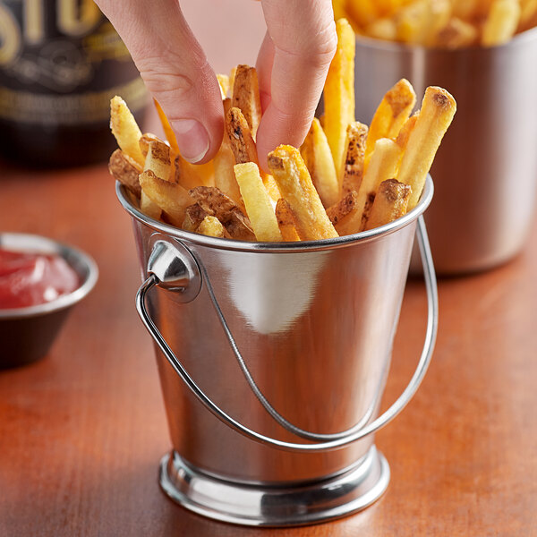 A person's finger holding a french fry and reaching into a Vollrath mini stainless steel bucket full of french fries.