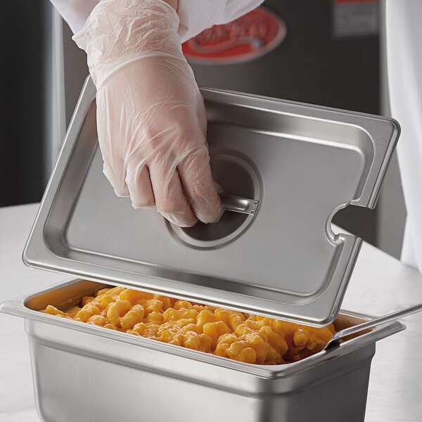 A person in plastic gloves holding a Carlisle stainless steel slotted steam table pan cover over a container of macaroni and cheese.