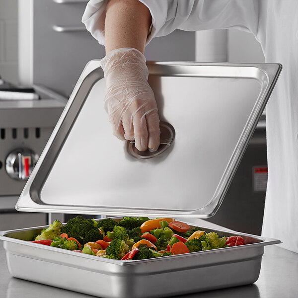 A person in a gloved hand holding a Carlisle stainless steel steam table pan cover filled with broccoli and tomatoes.