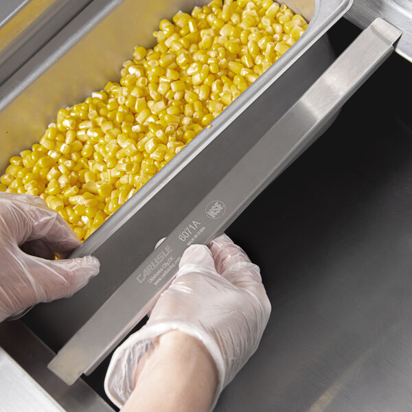 A person in gloves using a Carlisle stainless steel steam table pan adapter bar to hold a pan of corn.
