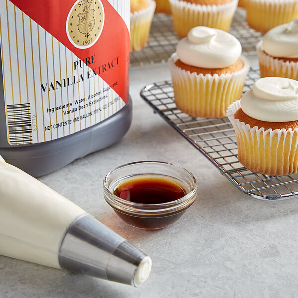 A cupcake with white frosting on top next to a bowl of brown liquid and a container of Nielsen-Massey Pure Vanilla Extract.