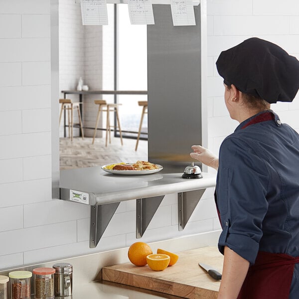 A woman in a blue uniform and chef's hat preparing food on a Regency stainless steel pass-through shelf.