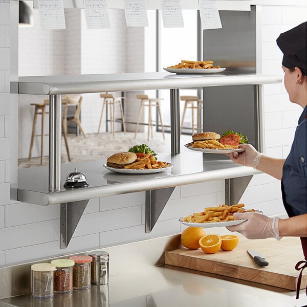 A woman in a chef's uniform in a school kitchen using a Regency stainless steel pass-through shelf to hold plates of food.