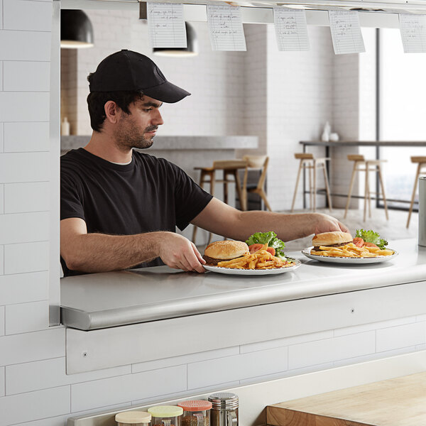 A man in a black hat sitting at a counter with plates of food on a Regency stainless steel pass-through shelf.