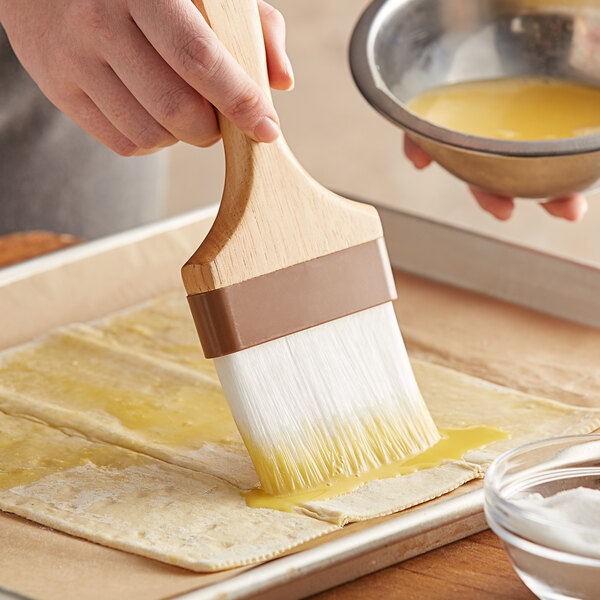 A hand using a Thunder Group nylon bristle pastry brush to spread yellow liquid over a pastry.