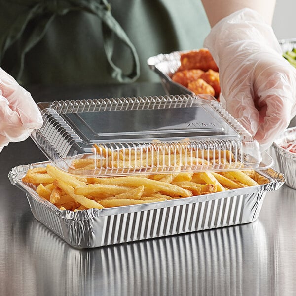 A gloved hand putting french fries in a Choice oblong foil take-out container.