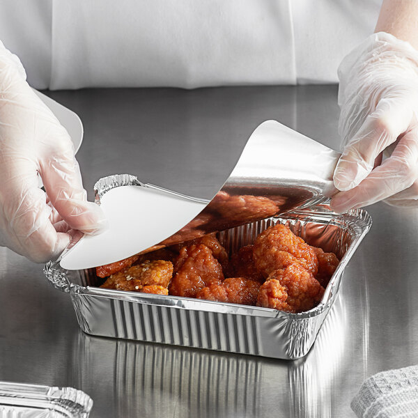 A person wearing a plastic glove putting chicken wings in a foil container with a Choice foil-laminated board lid.