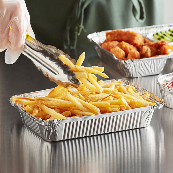 A person using tongs to put French fries in an oblong foil take-out container.