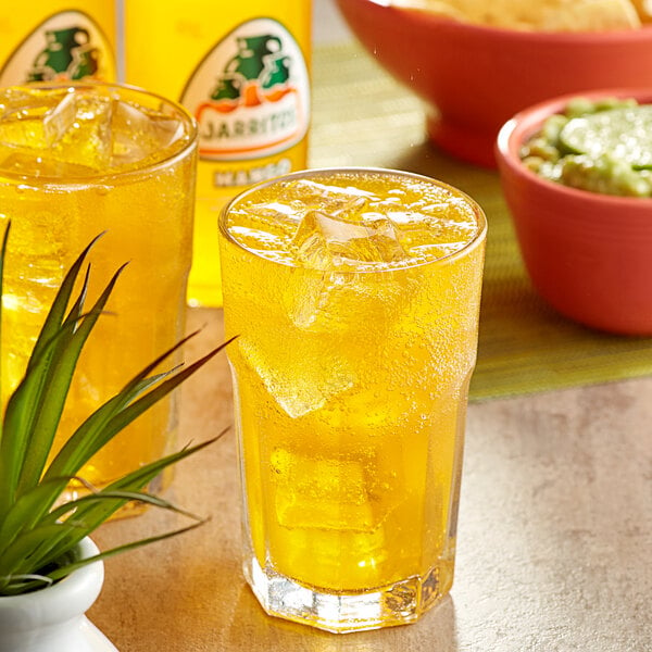 A glass of Jarritos Mango soda with ice cubes on a table with a bowl of food and a plant.