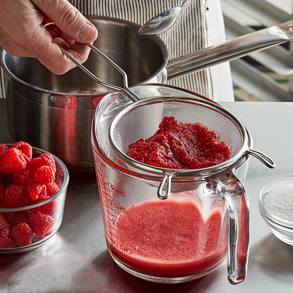 A hand using a Tablecraft fine mesh strainer to stir raspberries in a glass container.