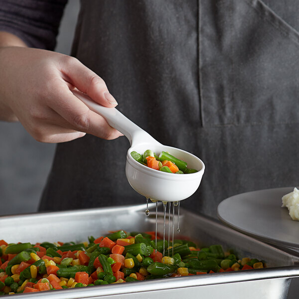 A hand using a Carlisle white perforated portion spoon to pour vegetables into a container.