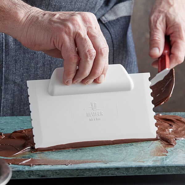 A hand using a Matfer Bourgeat chocolate scraper to decorate chocolate on a white object.