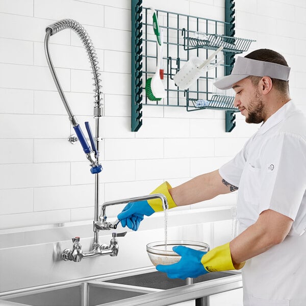A man in gloves washing dishes in a sink using a Waterloo wall-mounted pre-rinse faucet.