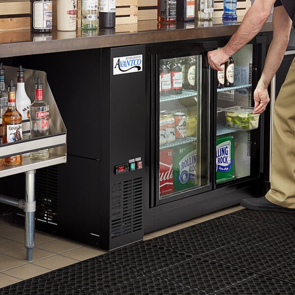 A man standing at a bar with a black Avantco back bar refrigerator full of drinks.