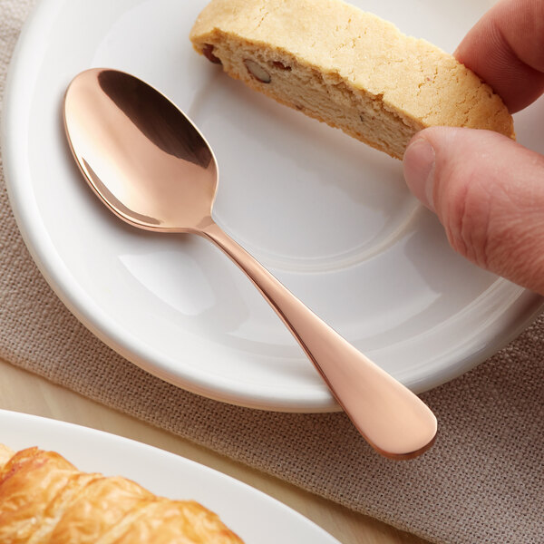 A hand holding a piece of pastry on a plate with an Acopa Vernon rose gold demitasse spoon.