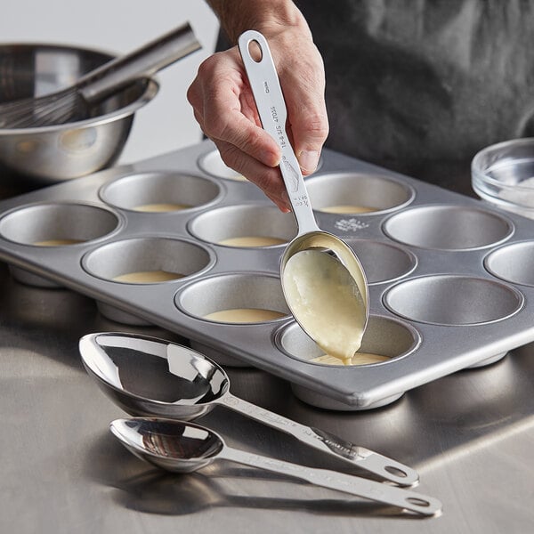 A hand using Vollrath stainless steel measuring spoons to scoop batter into a muffin tin.