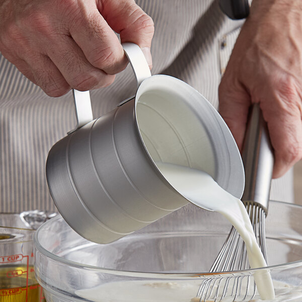 A person using a Vollrath aluminum measuring cup to pour milk into a bowl.