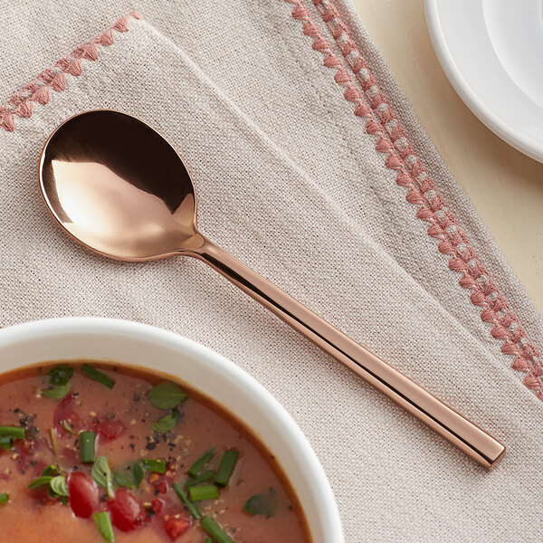 An Acopa Phoenix rose gold stainless steel bouillon spoon in a bowl of soup on a table with a napkin.