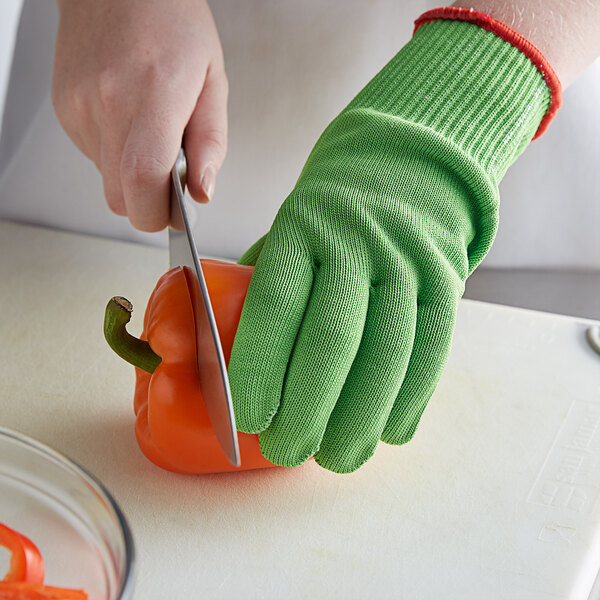 A person wearing Mercer Culinary green cut-resistant gloves cutting a red bell pepper on a counter.