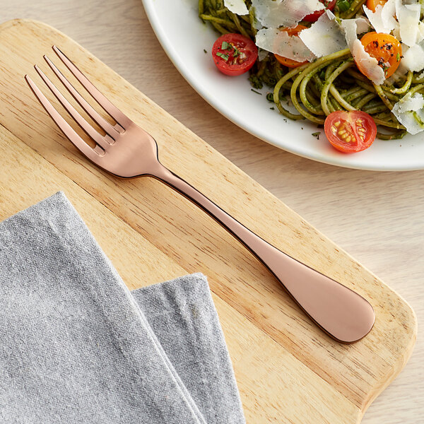 An Acopa Vernon rose gold stainless steel dinner fork on a cutting board next to a plate of pasta.