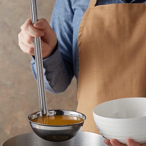 A person holding a Vollrath stainless steel ladle over a white bowl of brown liquid.
