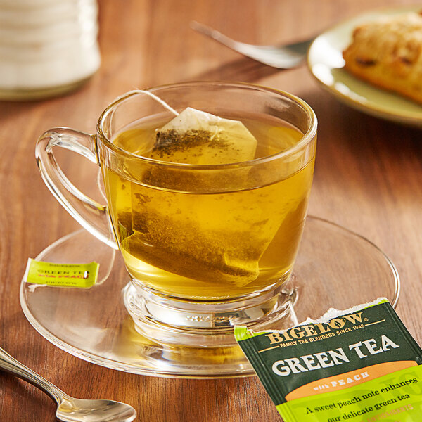 A glass cup of Bigelow Green Tea with a tea bag on a saucer with a spoon on the table.