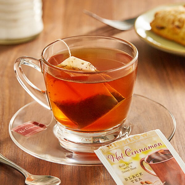 A glass cup of Bigelow Hot Cinnamon tea with a tea bag on a saucer.