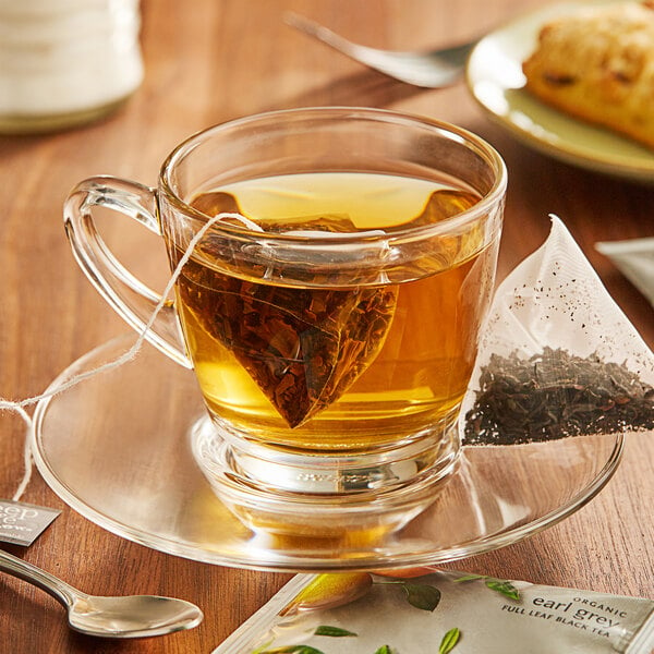 A glass cup of Steep Cafe By Bigelow Earl Grey tea with a tea bag on a saucer and a spoon.
