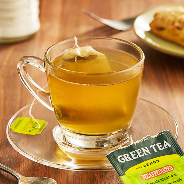 A glass cup of Bigelow Green Tea with Lemon Decaffeinated Tea on a saucer with a tea bag.