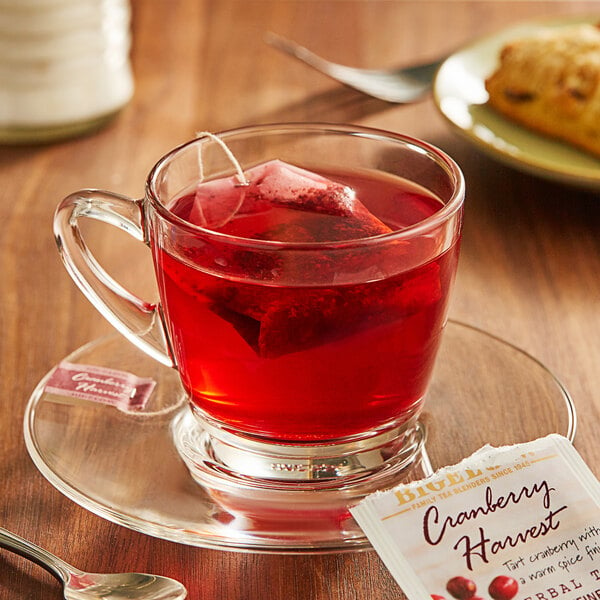 A glass cup of Bigelow Cranberry Harvest tea with a tea bag on a saucer.