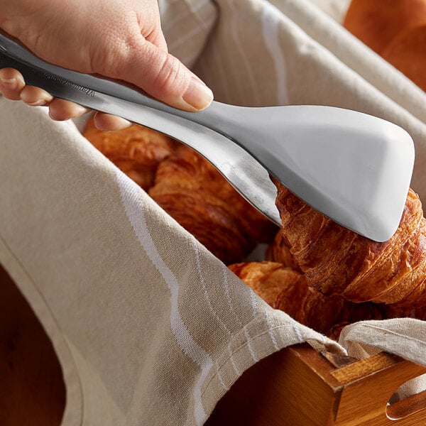 A hand using Vollrath stainless steel bread tongs to pick up a croissant.