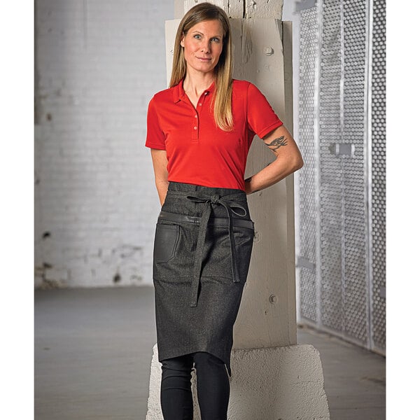 A woman wearing a red shirt and Mercer Culinary black denim bistro apron with black leather details standing next to a white wall.