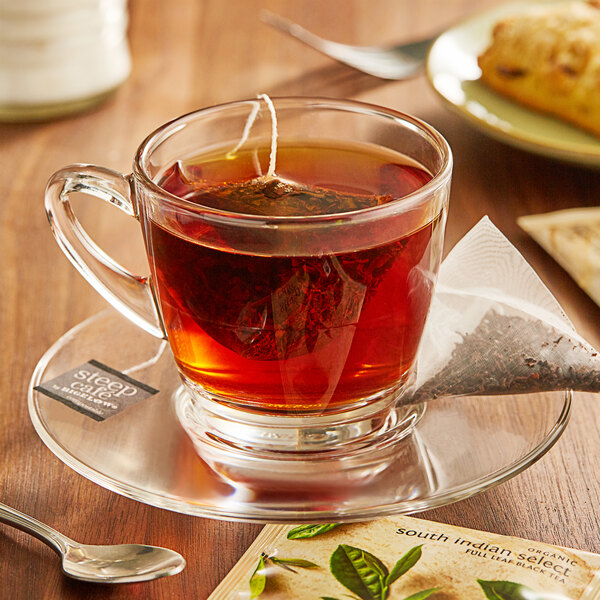 A glass cup of Steep Cafe By Bigelow Organic South Indian Select Black Tea with a tea bag on a saucer and a spoon.