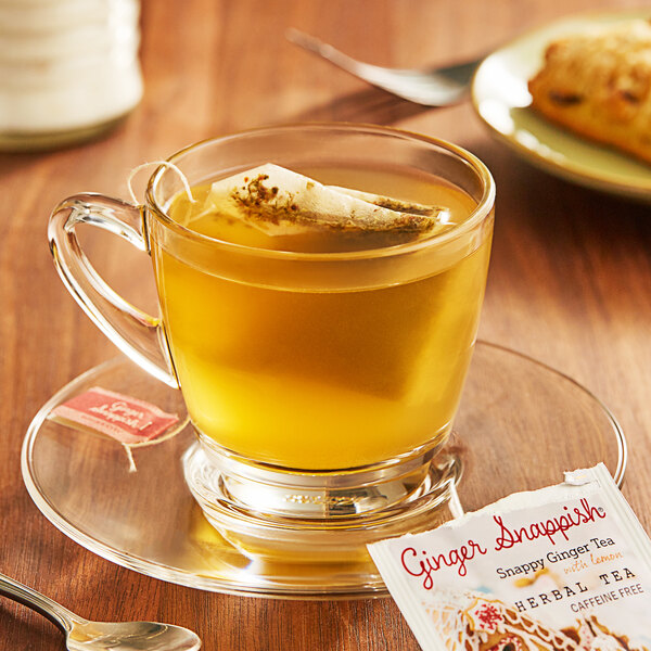 A glass cup of Bigelow Ginger Snappish Herbal Tea with a tea bag on a plate.
