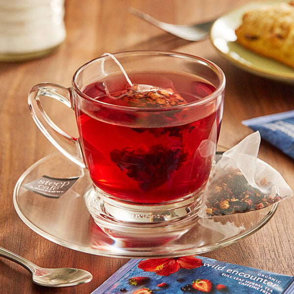 A glass cup of Steep Cafe wildberry hibiscus tea with a bag of tea on a saucer.