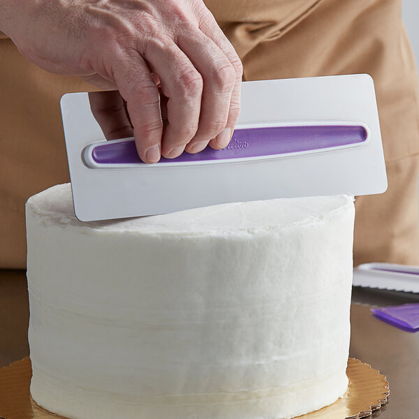 A hand using Wilton stainless steel icing comb to decorate a white cake.